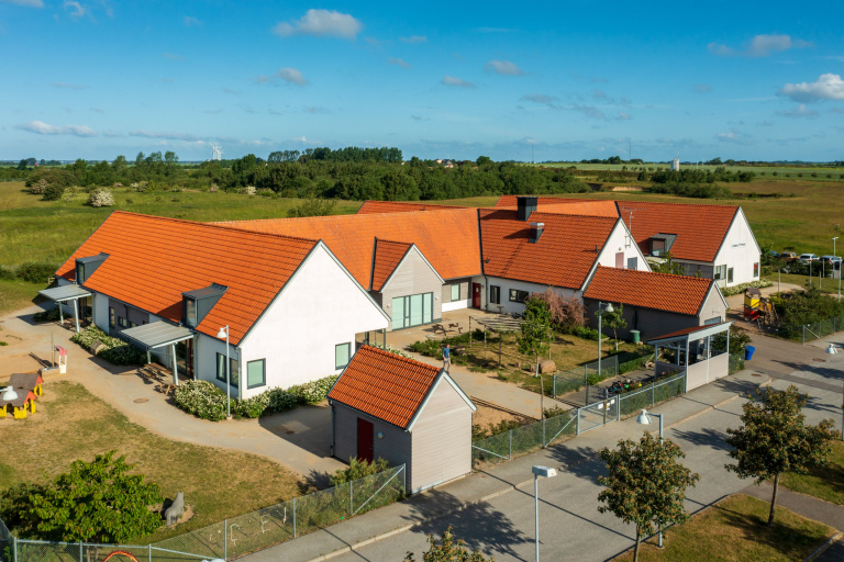 Vitt en och en halvplanshus med rött tak, byggt med utskjutande flyglar. Bilden r fotograferad från ovan och visar även delar av gård och lekplats.