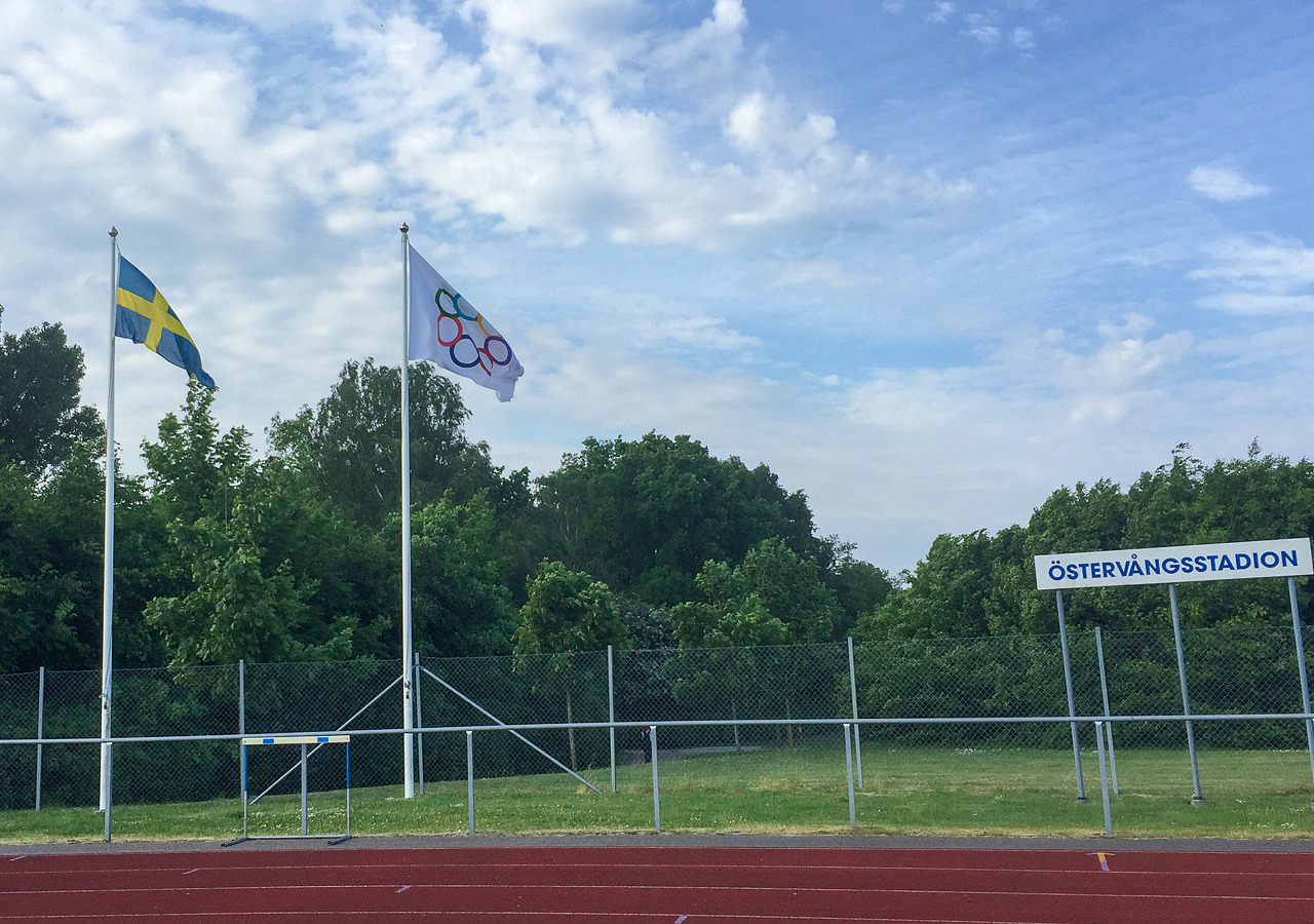 Två flaggstänger, en svensk flagga och en OS-flagga. Skylt med texten Östervångstadion.