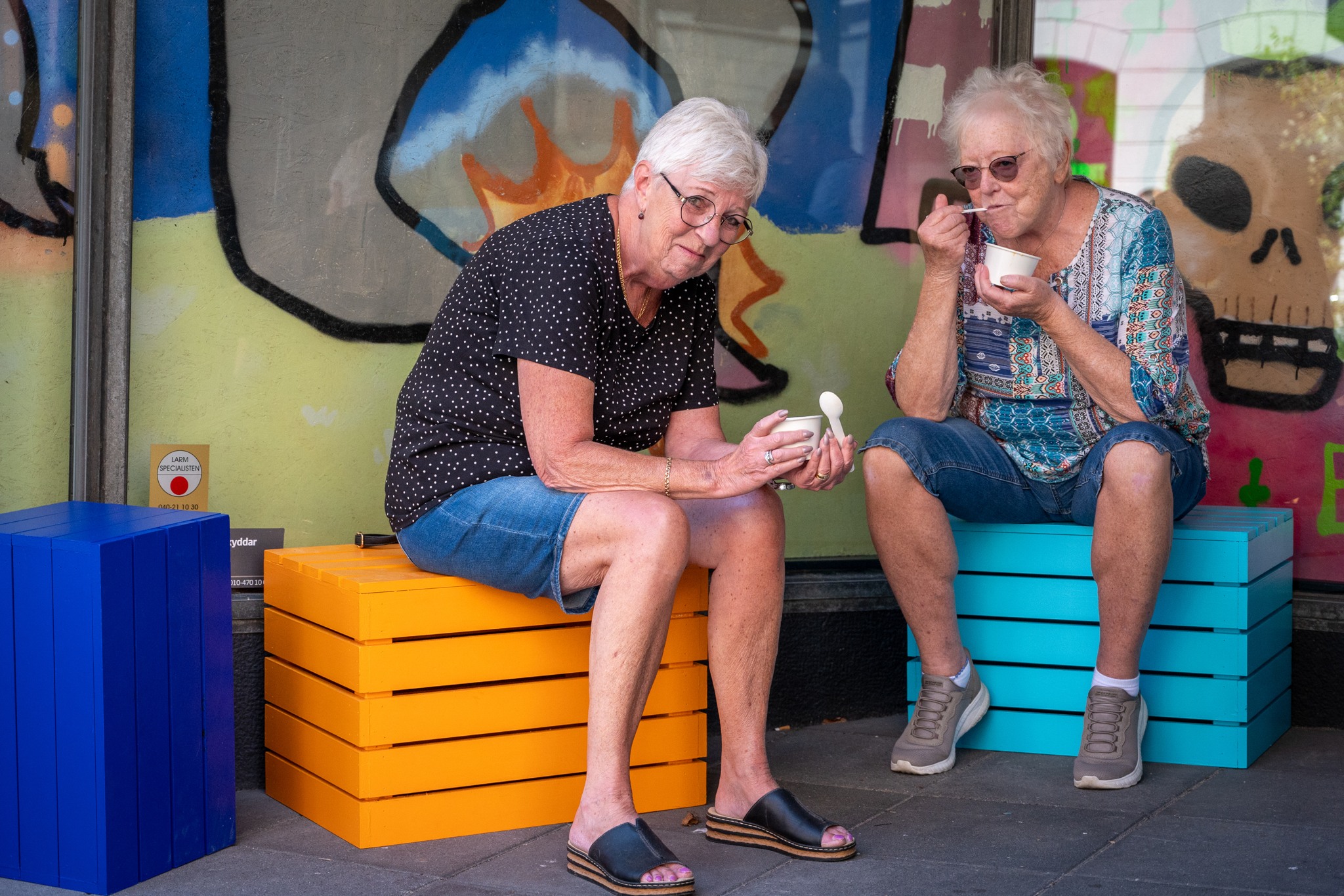 Två kvinnor sitter på färgglada pallar och äter mat.